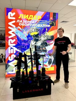 Мужчина указывает на стойку с оружием LASERWAR у баннера.