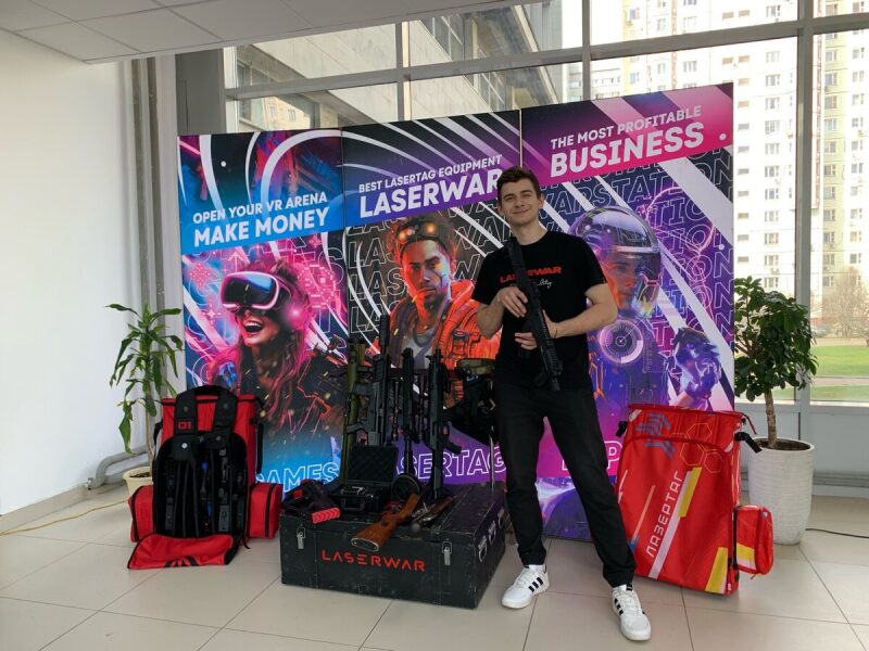 Молодой человек демонстрирует лазертаг-автомат у баннера LASERWAR.