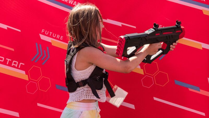 Девочка в жилете LASERWAR целится из футуристического автомата.