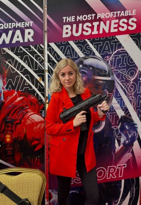 Женщина в красном пиджаке держит лазертаг-пистолет LASERWAR.